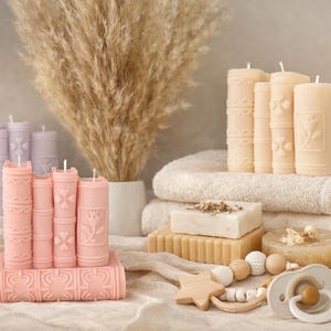 May include: A collection of decorative candles shaped like books in shades of pink and cream. The candles are arranged with other items, including soap, towels, and a baby pacifier. The candles have detailed designs and are a unique home decor item.
