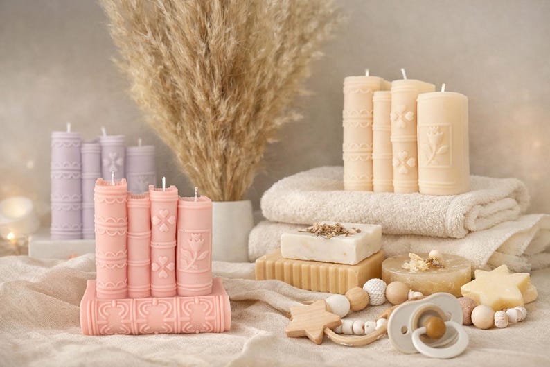 May include: A collection of decorative candles shaped like books in shades of pink and cream. The candles are arranged with other items, including soap, towels, and a baby pacifier. The candles have detailed designs and are a unique home decor item.