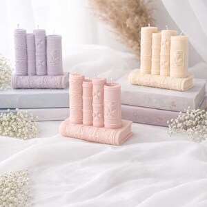 May include: Three sets of decorative candles shaped like books. The candles are in pastel colors: lavender, peach, and light yellow. Each set has a base shaped like a book with the candles arranged on top. The candles have floral and scrollwork details.