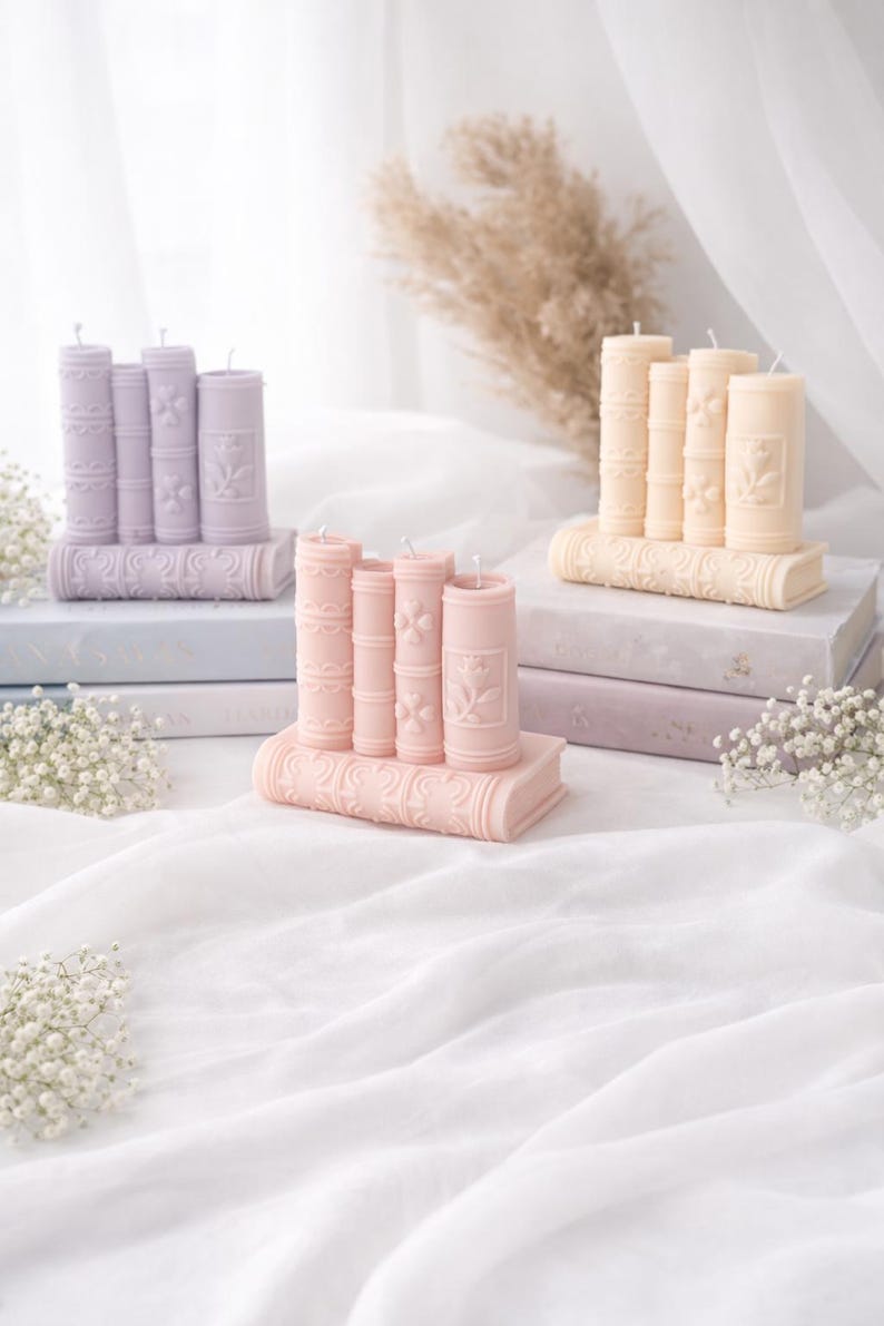 May include: Three sets of decorative candles shaped like books. The candles are in pastel colors: lavender, peach, and light yellow. Each set has a base shaped like a book with the candles arranged on top. The candles have floral and scrollwork details.