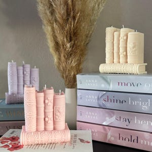May include: A display of decorative candles shaped like books in various pastel colors. The candles are arranged on top of and next to a stack of books with titles like "move on" and "shine bright". A vase of dried pampas grass is in the background.