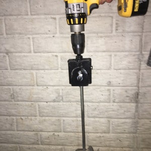 May include: A yellow and black cordless drill with a black mixing attachment and a silver mixing paddle. The drill is attached to a black metal bracket mounted on a white brick wall. The drill is labeled "DEWALT 18V"
