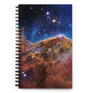 Op de afbeelding: Een spiraalgebonden notitieboek met een kleurrijk beeld van een nevel in de ruimte. De nevel is een mix van rode, oranje en blauwe kleuren met heldere witte sterren.