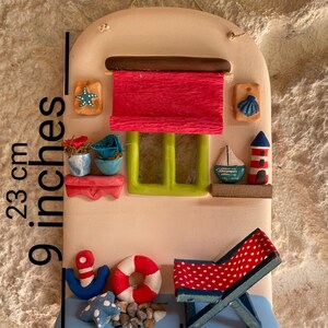 Puede incluir: Una decoraci&oacute;n de pared de cer&aacute;mica con forma de casita con un toldo rojo, una ventana verde y una silla de playa a rayas azules y blancas. La casa tiene un ancla azul y blanca, un salvavidas rojo y una estrella de mar en la parte inferior. La decoraci&oacute;n de pared mide 15 cm de ancho y 58 cm de alto.