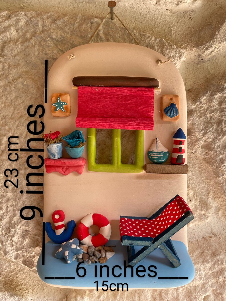 Puede incluir: Una decoraci&oacute;n de pared de cer&aacute;mica con forma de casita con un toldo rojo, una ventana verde y una silla de playa a rayas azules y blancas. La casa tiene un ancla azul y blanca, un salvavidas rojo y una estrella de mar en la parte inferior. La decoraci&oacute;n de pared mide 15 cm de ancho y 58 cm de alto.