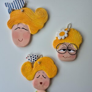 Può includere: Tre ornamenti a forma di viso in ceramica con capelli gialli, volti dipinti e dettagli decorativi. Uno ha un fiocco a righe blu e bianche, un altro una margherita e il terzo un uccello a pois. Ognuno ha un anello per appenderlo.
