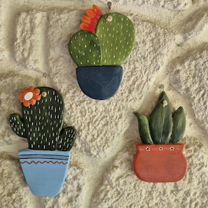 Pode incluir: Três decorações de parede de cactos de madeira. Cada cacto é pintado de verde com detalhes brancos e está em um vaso colorido. Um tem uma flor laranja, outro uma flor vermelha e o terceiro não tem flor. Os vasos são azuis e vermelhos.