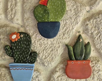 May include: Three wooden cactus wall hangings. Each cactus is painted green with white accents and sits in a colorful pot. One has an orange flower, another has a red flower, and the third has no flower. The pots are blue and red.