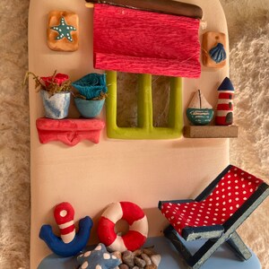 Puede incluir: Una decoraci&oacute;n de pared de cer&aacute;mica que representa una caba&ntilde;a costera con un toldo rojo, una ventana con contraventanas verdes y un ancla azul. La caba&ntilde;a tiene un peque&ntilde;o balc&oacute;n con dos plantas en macetas. Hay un salvavidas rojo y blanco, una estrella de mar azul y blanca y una silla de playa roja y blanca con lunares blancos.