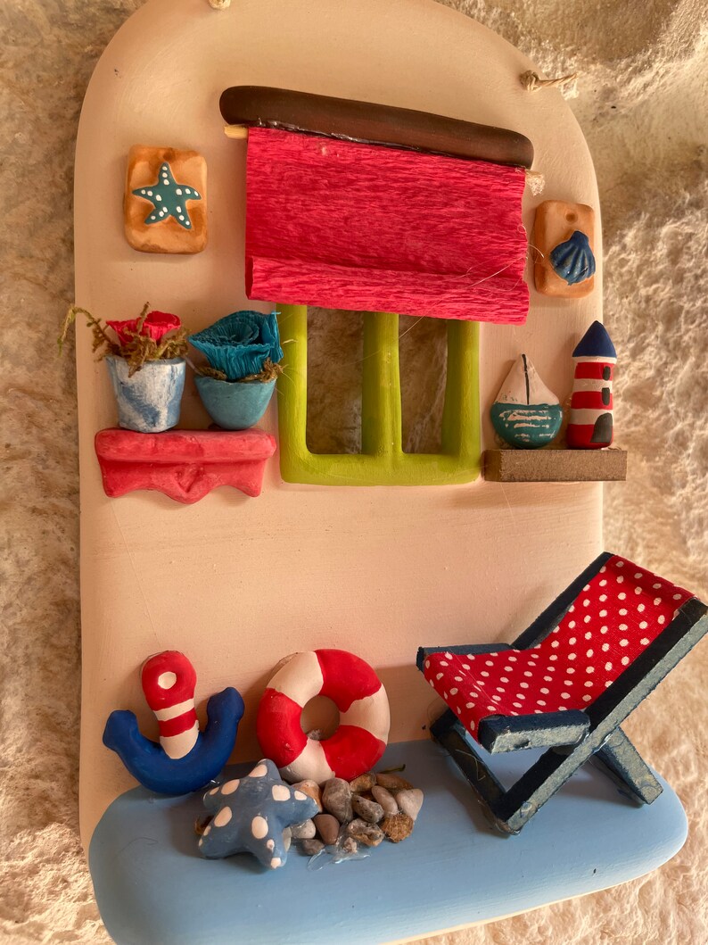 Puede incluir: Una decoraci&oacute;n de pared de cer&aacute;mica que representa una caba&ntilde;a costera con un toldo rojo, una ventana con contraventanas verdes y un ancla azul. La caba&ntilde;a tiene un peque&ntilde;o balc&oacute;n con dos plantas en macetas. Hay un salvavidas rojo y blanco, una estrella de mar azul y blanca y una silla de playa roja y blanca con lunares blancos.