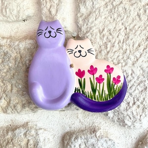 May include: A whimsical cat-themed wall hanging. One cat is lavender, the other is light pink with a purple tail. The pink cat features a floral design with pink flowers and green stems. The cats are attached and mounted on a white brick wall.