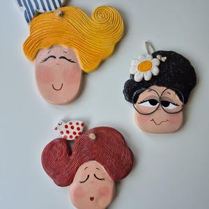 Peut inclure: Trois ornements de visage en céramique fantaisistes. L'un a des cheveux jaunes et un nœud rayé bleu et blanc. Un autre a des cheveux noirs et des lunettes avec une marguerite. Le troisième a des cheveux roux et un oiseau à pois rouges.