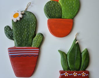 May include: Three wooden cactus and succulent wall hangings. Each plant is painted green and sits in a red pot. One cactus has a white flower, another has an orange flower, and the third has green leaves.
