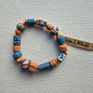 Könnte beinhalten: Ein Armband aus terrakottafarbenen Tonperlen und blau bemalten Keramikperlen mit weißen Blatt- und Herzmustern, getrennt durch kleine silberfarbene Metallabstandshalter. Ein braunes Papieretikett mit der handschriftlichen Aufschrift Hand made ist befestigt.