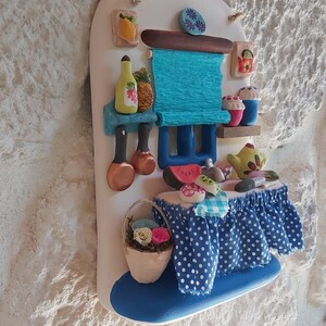 May include: A white ceramic wall hanging with a blue and white polka dot tablecloth, a blue and white striped awning, and a variety of colorful ceramic food items, including a watermelon, a pineapple, and a teapot.