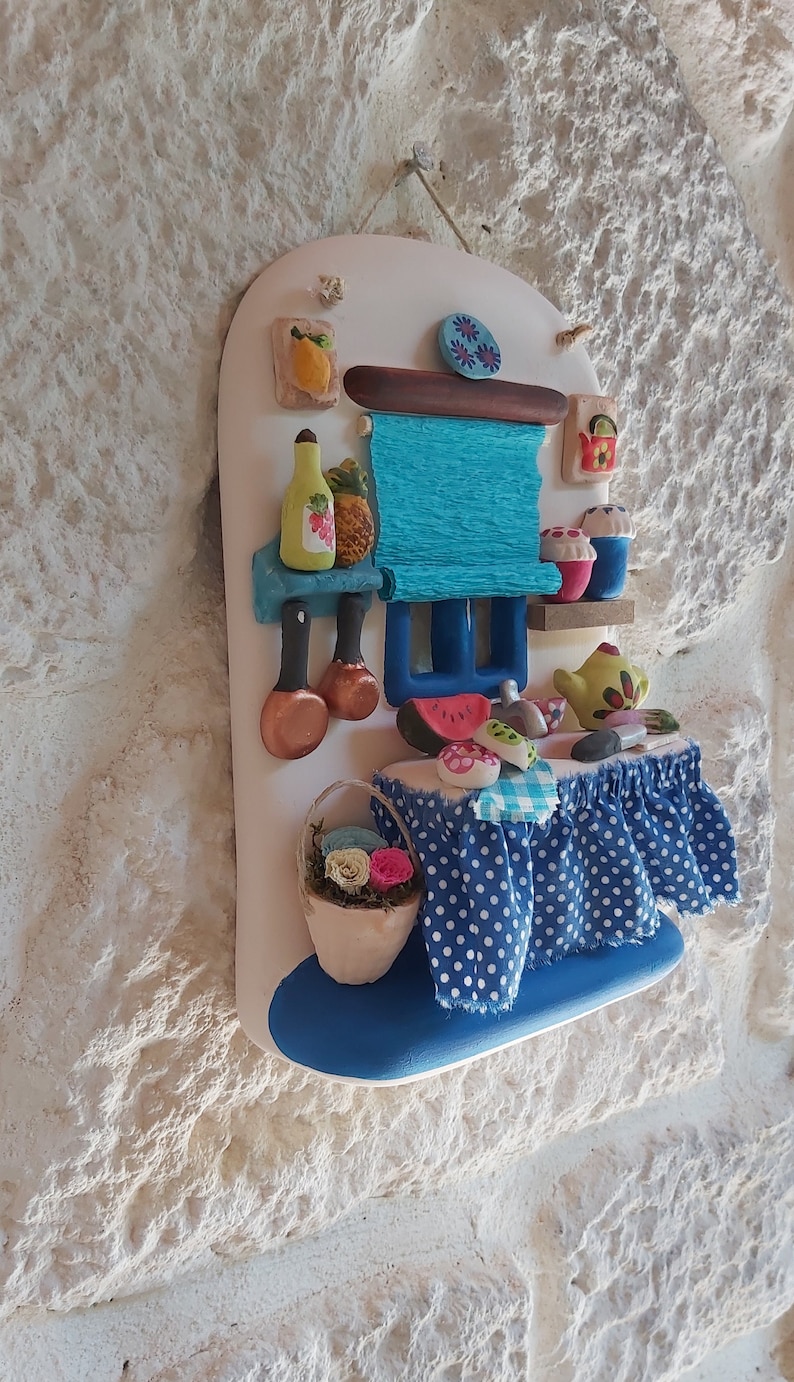 May include: A white ceramic wall hanging with a blue and white polka dot tablecloth, a blue and white striped awning, and a variety of colorful ceramic food items, including a watermelon, a pineapple, and a teapot.
