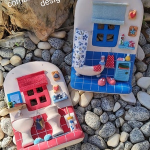 May include: Two ceramic bathroom-themed miniature houses with red and blue tile details. The house on the left has a red roof and the house on the right has a blue roof. Both houses have white walls and windows. The houses are sitting on a bed of white and gray stones.