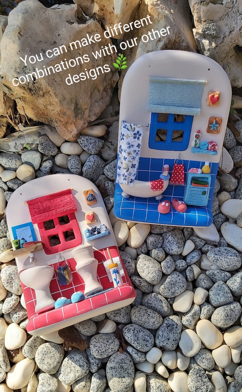 May include: Two ceramic bathroom-themed miniature houses with red and blue tile details. The house on the left has a red roof and the house on the right has a blue roof. Both houses have white walls and windows. The houses are sitting on a bed of white and gray stones.