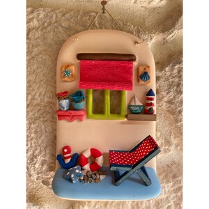 Puede incluir: Una decoraci&oacute;n de pared de cer&aacute;mica con forma de casita con un toldo rojo, una ventana con contraventanas verdes y una silla de playa a rayas azules y blancas. La casa tiene un toldo a rayas azules y blancas y una silla de playa a rayas azules y blancas. Tambi&eacute;n hay peque&ntilde;os detalles decorativos como una estrella de mar, una concha y un faro.