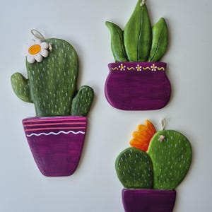 May include: Three wooden cactus decorations in purple pots. The cacti are green with white accents, and one has a daisy, another has a yellow flower, and the third has green leaves. Each has a string for hanging.
