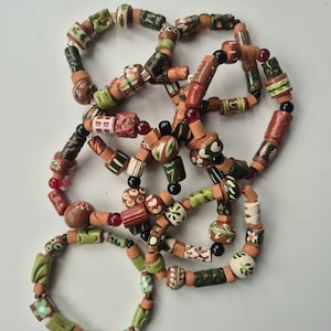 Könnte beinhalten: Eine Sammlung handgefertigter Armbänder mit terrakottafarbenen Tonperlen, die mit kleinen, runden, durchscheinenden roten und schwarzen Glasperlen durchsetzt sind. Die Tonperlen sind mit handgemalten grünen, weißen und braunen Pflanzenmotiven verziert.