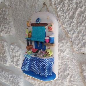May include: A white ceramic wall hanging depicting a miniature kitchen scene. The kitchen features a blue and white checkered tablecloth, a blue sink, a watermelon, and a green teapot.