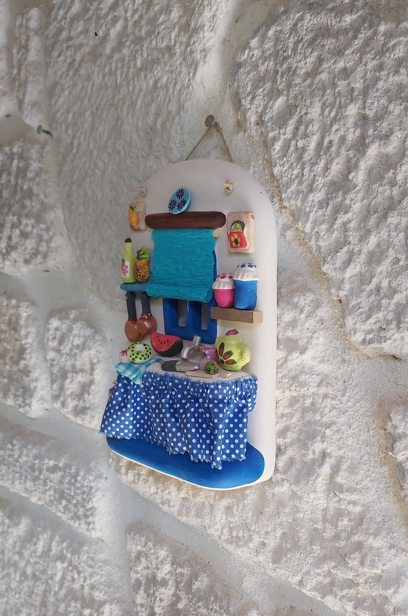 May include: A white ceramic wall hanging depicting a miniature kitchen scene. The kitchen features a blue and white checkered tablecloth, a blue sink, a watermelon, and a green teapot.