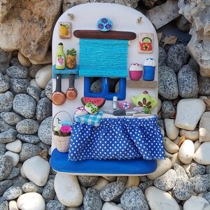 May include: A miniature kitchen scene with a blue and white polka dot tablecloth, a blue window with a watermelon slice, and a blue awning. The kitchen is decorated with painted details, including a teapot, a vase of flowers, and a pineapple.