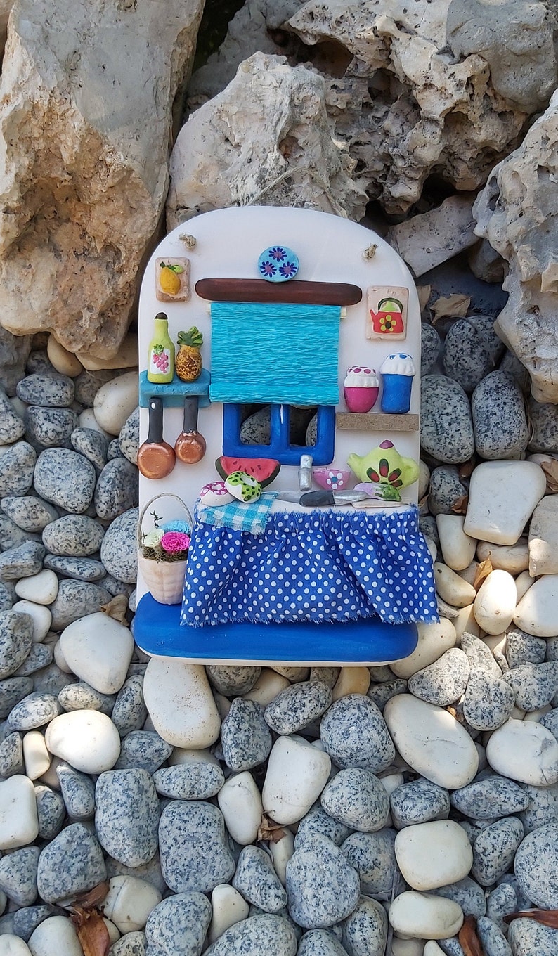 May include: A miniature kitchen scene with a blue and white polka dot tablecloth, a blue window with a watermelon slice, and a blue awning. The kitchen is decorated with painted details, including a teapot, a vase of flowers, and a pineapple.