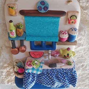 May include: A ceramic kitchen scene with a blue and white checkered tablecloth, a watermelon, two donuts, a teapot, a red kettle, and a blue and white striped window blind.