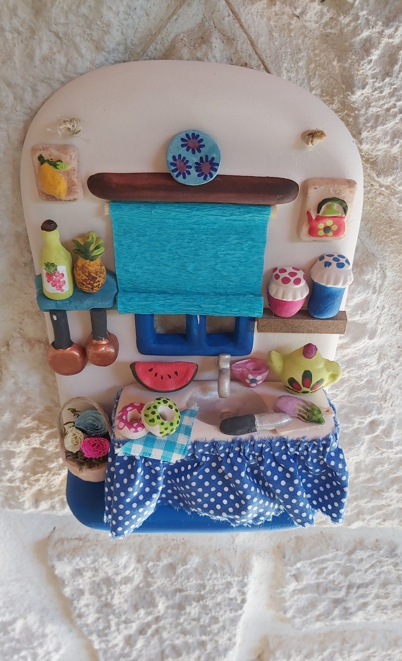 May include: A ceramic kitchen scene with a blue and white checkered tablecloth, a watermelon, two donuts, a teapot, a red kettle, and a blue and white striped window blind.