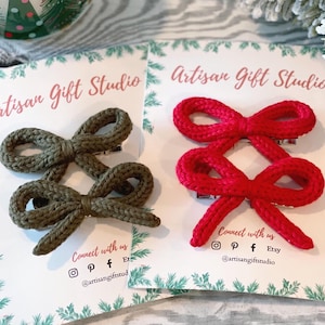 Puede incluir: Dos juegos de lazos para el pelo tejidos a mano. Un juego es de color verde oliva y el otro es de color rosa brillante. Ambos juegos se muestran en tarjetas blancas con el texto "Artisan Gift Studio" y la información de contacto de las redes sociales.