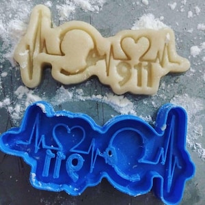 Pode incluir: Um biscoito de cor clara em forma de monitor de frequência cardíaca médica com os números "911" e um símbolo de coração. Um cortador de biscoito de plástico azul com a palavra "fire" está abaixo.
