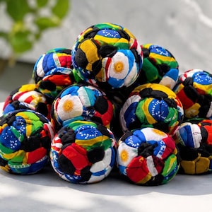 May include: A collection of colorful, patchwork-style soccer balls. Each ball is made of fabric pieces in various colors and patterns, including flags of different countries. The balls are stacked together, creating a vibrant and playful display.