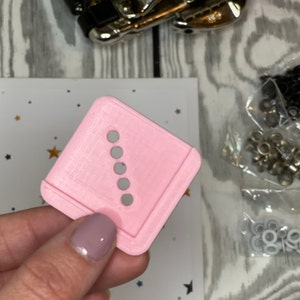 May include: Pink plastic tool with six evenly spaced holes in a vertical line. The tool is used for setting eyelets.