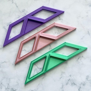 May include: Three geometric, diamond-shaped objects in purple, pink, and green. Each object has a rectangular opening and a textured surface. The objects are arranged diagonally on a white and grey surface.