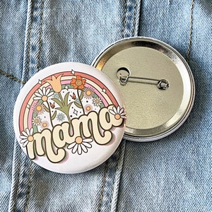 May include: A white button with a rainbow, flowers, and the word "mama" in a retro font. The button is attached to a silver pin back.