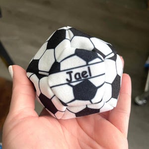 May include: A white and black soccer ball-shaped item with the name "Jael" printed on it. The item is held in a hand. The soccer ball design features black pentagons and white hexagons.