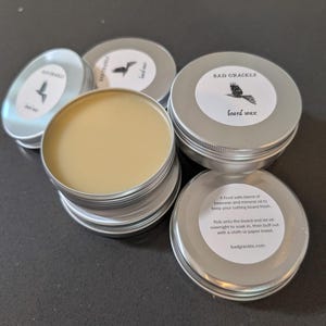 Puede incluir: Varios botes pequeños de cera para tablas de color plateado, con etiquetas blancas con un gráfico de un pájaro negro y el texto "BAD GRACKLE board wax". Un bote está abierto, revelando una cera de color amarillo claro.