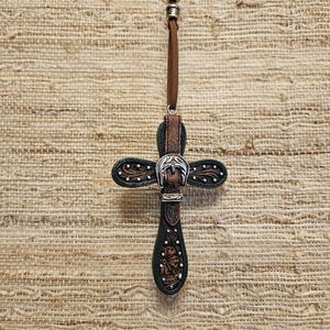 May include: A decorative cross pendant with a western-style design. The cross is made of dark brown and black leather, with silver-colored metal accents. The pendant hangs from a brown cord with silver beads.