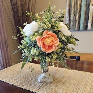 May include: A floral centerpiece with peach and white flowers arranged in a white pedestal vase. The flowers are surrounded by green foliage and are displayed on a beige table runner.