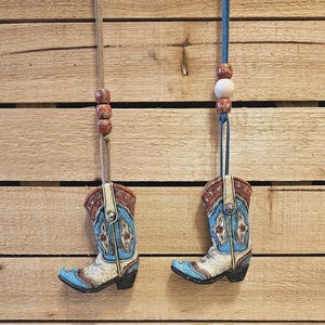 May include: Two blue and white cowboy boot ornaments hanging from brown and blue cords with wooden beads. The boots have red and white accents and are hanging against a wood background.