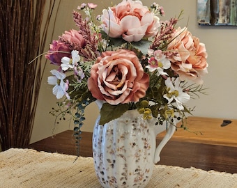 May include: A white ceramic pitcher with a ribbed texture and a delicate brown floral pattern, filled with an arrangement of artificial flowers including large dusty rose peonies, smaller white blossoms, and various green foliage.