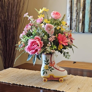 May include: A ceramic roller skate planter in white with red and yellow accents, featuring a painted yellow flower on the side. The skate is filled with an arrangement of artificial pink peonies, yellow wildflowers, and various green foliage.