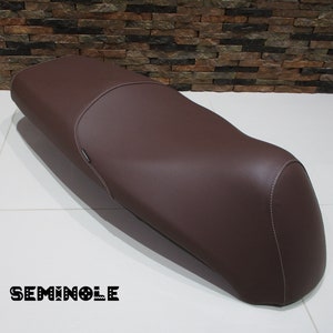 May include: A brown leather motorcycle seat with white stitching. The seat is curved and has a slight hump in the middle. The word "SEMINOLE" is printed on the floor in front of the seat.