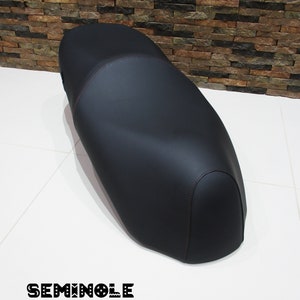 May include: Black leather motorcycle seat with brown stitching. The seat is on a white surface with a brick wall in the background.