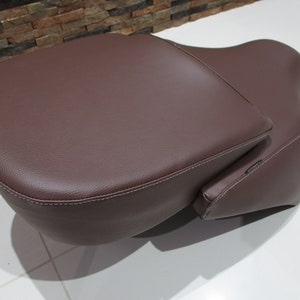 May include: A brown leather motorcycle seat with a passenger backrest. The seat has a stitched edge and a smooth surface.