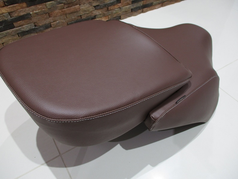 May include: A brown leather motorcycle seat with a passenger backrest. The seat has a stitched edge and a smooth surface.