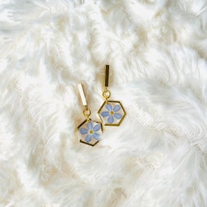 May include: A pair of gold-toned earrings featuring a hexagonal frame with a delicate blue flower encased within. The earrings have a rectangular top piece and hang from a small gold loop. The background is a soft, white, fluffy texture.