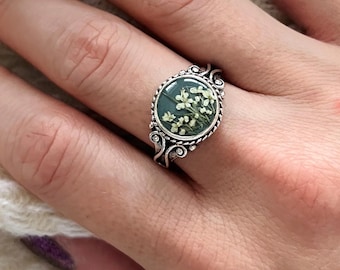 Puede incluir: Un anillo de plata con un engaste circular que contiene un fondo verde oscuro y pequeñas flores blancas prensadas encapsuladas en resina. La banda presenta una textura de cuerda trenzada y detalles metálicos ornamentados en los laterales.
